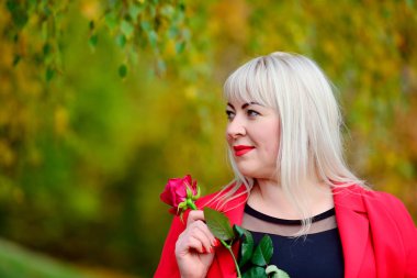 Kırmızı bir ceket ve bir kızıl bir sarışın büyük bir portre elinde bir güneşli sonbahar gününde bir parkta gül. Hayal kurmak ve uzağa bakmak. Kadın büyüklüğü artı XXL. Yatay görünüm