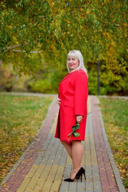 Kırmızı bir ceket güzel bir büyük sarışın ve elinde bir kızıl gül bir güneşli sonbahar gününde Park bir yolda pozlar. Kadın büyüklüğü artı XXL. Genel plan. Dikey görünüm