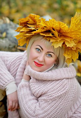Sonbahar yaprakları bir çelenk güzel sarışın, sarı bir arka planda ormanda nazik bir Jumper oturan, kamera bakarak ve gülümseyen. Kadın büyüklüğü artı XXL