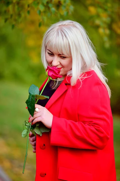 A beautiful large blonde in a black dress and a red coat bent thoughtfully over the rose in her hands. Sunny autumn day. Woman's size plus xxl. Close up. Vertical view