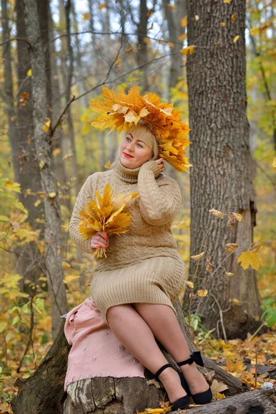 A sweet woman, a middle-aged blonde in autumn leaves, with a wreath on her head, sitting on a stump in the autumn forest in a gentle jumper. Size plus xxl female. Vertical view