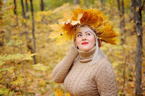 Приваблива жінка, блондинка середнього віку в осінньому листі, з вінком на голові в осінньому лісі в бежевому джемпері. Вона задумливо дивиться вгору. Розмір плюс Xxl жіночий. Горизонтальний вигляд — стокове фото