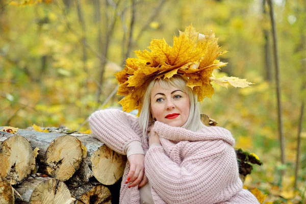 가을 단풍화환의 아름다운 금발, 나무통에 기대어 숲에서 부드러운 점퍼에 앉아, 신중하고 미소. 여성 사이즈 플러스 xxl — 스톡 사진