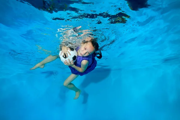 Cute girl swimming and playing underwater with the ball. On a blue background in a dress, eyes open. Portrait. Horizontal view