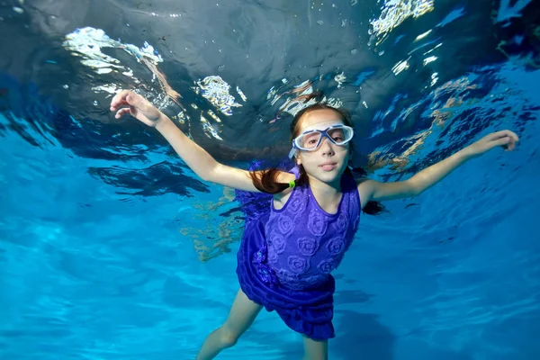 Gelukkig klein meisje zwemmen onderwater in het zwembad. Ze glimlacht en kijkt naar de camera in een paarse jurk en zwembrillen, armen uitgerekt. Portret. Horizontale weergave — Stockfoto