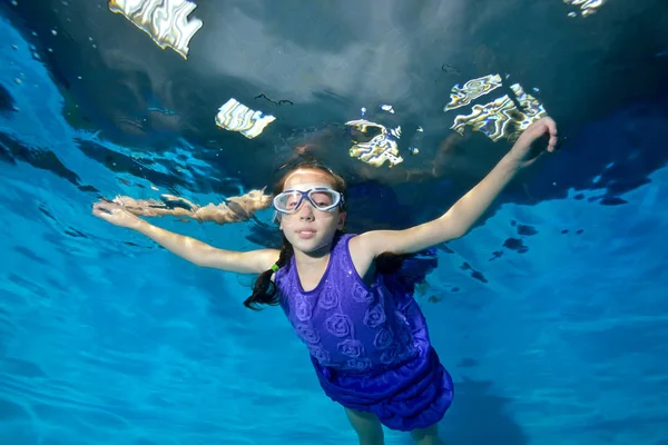 Gelukkig, lachend kind. Een meisje in een paarse jurk en zwembrillen zwemt in het zwembad armen uitgestrekt. Onderwater fotografie. Horizontale weergave — Stockfoto