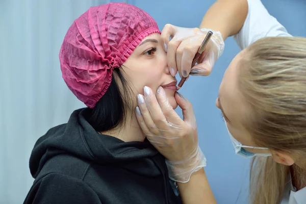 El esteticista con una bata blanca prepara al paciente para el procedimiento de los labios permanentes del maquillaje en el salón de belleza. Dibuja un dibujo a lápiz en los labios de una chica sentada. Retrato. Vista horizontal — Foto de Stock