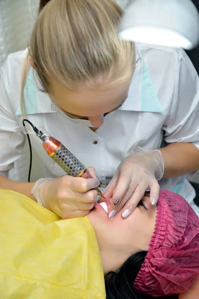 Mujer-cosmetóloga durante el procedimiento de los labios de maquillaje permanente, que hace chica europea acostada en el sofá en el salón de belleza. Concepto. Orientación vertical de la imagen — Foto de Stock