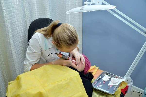 A master beautician, blonde, in a white medical gown makes permanent lip makeup to a young girl in a beauty salon and puts pink paint on her lips with a needle. Portrait. Horizontal view