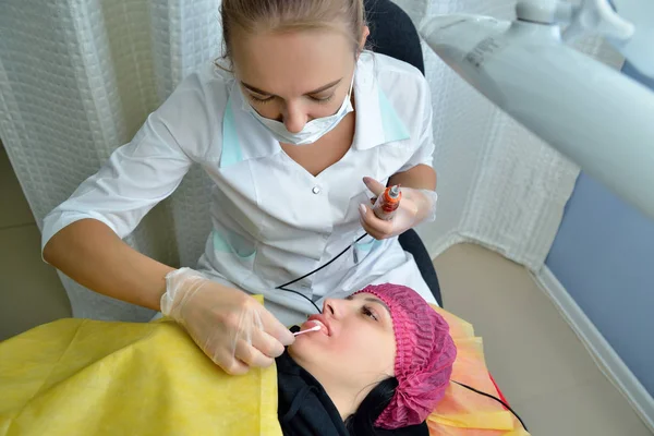 Kosmetolog, blond, sprawia, że Makijaż permanentny warg Europejskiej dziewczyny, robi połysk na ustach podczas zabiegu. Z bliska. Orientacja pozioma obrazu — Zdjęcie stockowe