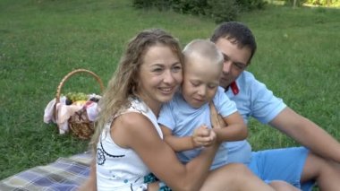 Sevimli aile - anne ve baba ve onların küçük oğlu frolic gün batımında Park'ta çim üzerinde oturan. Gülerler ve sarılırlar. Açık havada aile pikniği. L. Portre kavramı. 4k.