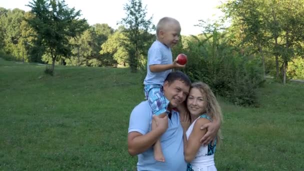 Mały chłopiec siedzi na ramionach ojca i trzyma jabłko w rękach. Mama stoi i ogodzi męża i syna. Z bliska. 4K. — Wideo stockowe