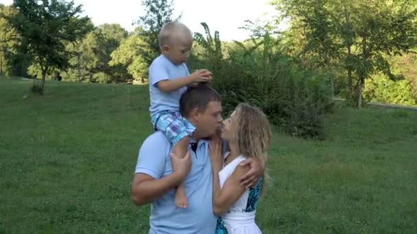 Gelukkige familie tijdens het wandelen in het Park in de zomer. Een jongetje zit op de schouders van zijn vader en houdt een appel in zijn handen. Hij speelt met papa en zal hem niet laten kus moeder. — Stockvideo