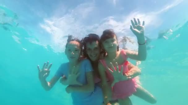 Família feliz: mãe e duas filhas, nadando e abraçando debaixo d 'água na piscina em um dia ensolarado. Eles posam para a câmera, sorriem e acenam as mãos. Close-up. Vista inferior. Movimento lento. 4K — Vídeo de Stock