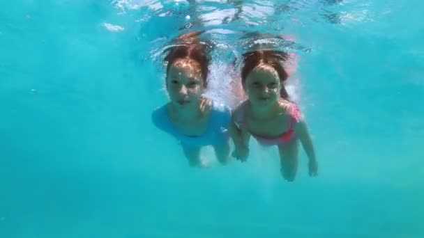 Due bambine felici nuotano sott'acqua tenendosi per mano in un costume da bagno rosa e blu in una giornata di sole. Guardano la telecamera e sorridono contro il cielo. Primo piano. Al rallentatore. 4K — Video Stock