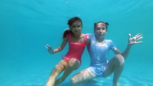 Duas garotinhas fofas abraçam e posam para a câmera subaquática no fundo da piscina em um maiô rosa e azul em um dia ensolarado. Eles olham para a câmera, sorriem e acenam as mãos. Movimento lento. 4K — Vídeo de Stock