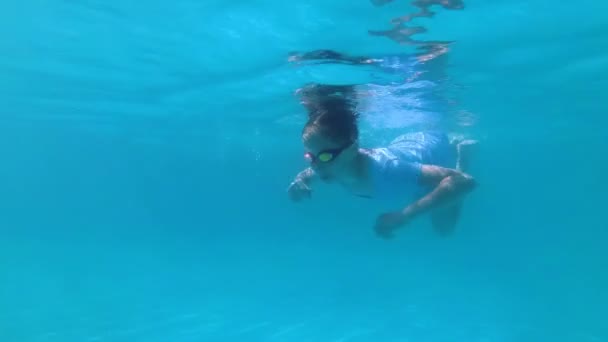 Una joven dulce se dedica a los deportes en una piscina al aire libre y nada bajo el agua en un traje de baño azul y gafas rojas para nadar en un día soleado. De cerca. En cámara lenta. 4K — Vídeo de stock