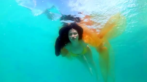 Egy gyönyörű lány egy sárga fürdőruhát úszik, és játszik a víz alatt egy sárga ruhával a kezében egy napsütéses napon. Ő pózok a medencében a haját, és nézi a kamerát. Lassított mozgás. — Stock videók