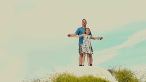 Papá feliz y su hija pequeña están de pie en una colina en un día soleado contra un cielo brillante. Se toman de la mano y comienzan a descender. El concepto de una familia feliz. 4K . — Vídeos de Stock