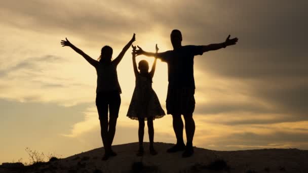 Papa, maman et leur petite fille sautent et agitent leurs bras contre le ciel jaune vif du coucher du soleil au sommet de la colline. Les parents embrassent la fille. Le concept d'une famille heureuse. 4K — Video