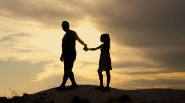 Babam ve küçük kızı tepenin tepesinden dramatik gün batımı gökyüzüne doğru inerler. Babam kızlarının elini tutuyor. Mutlu bir aile kavramı. Siluet. 4k