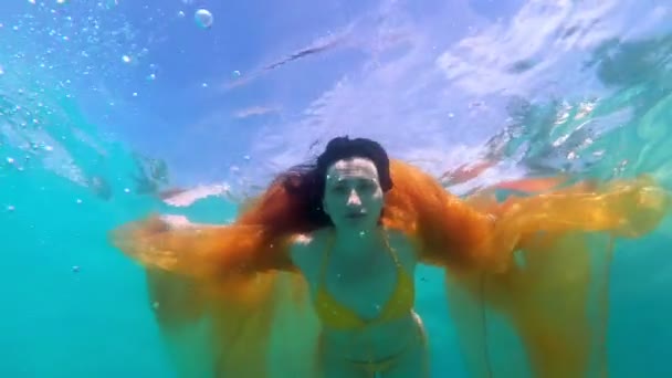 Een sprookjes meisje duikt onder het water met een gele doek in haar handen, in een geel badpak en zwemt naar de camera met haar haar op een zonnige dag. Close-up. Slow Motion. 4k. — Stockvideo