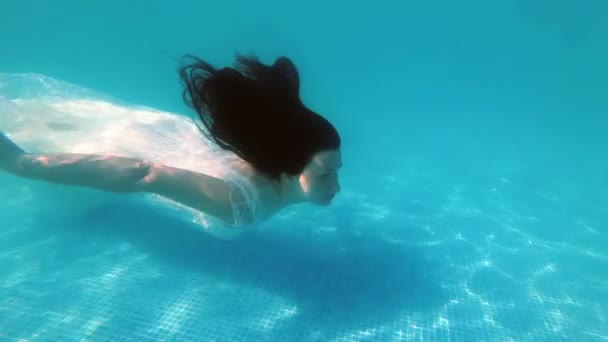 Une belle fille, une mariée en robe blanche, nage devant la caméra sous l'eau près du fond de la piscine les cheveux baissés par une journée ensoleillée. Gros plan. Au ralenti. 4K — Video