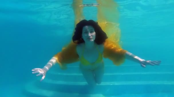Una chica de ensueño nada y posa delante de la cámara bajo el agua en la piscina en un traje de baño amarillo, con un paño amarillo en las manos y con el pelo suelto en un día soleado. En cámara lenta. 4K . — Vídeos de Stock