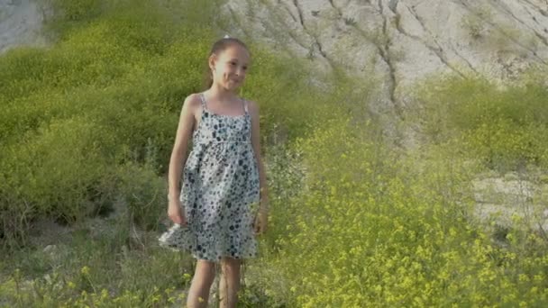 Una niña en un día soleado sobre el fondo de una montaña de tiza blanca se encuentra cerca de los arbustos de flores silvestres, sueña y agita suavemente su vestido. 4K — Vídeo de stock