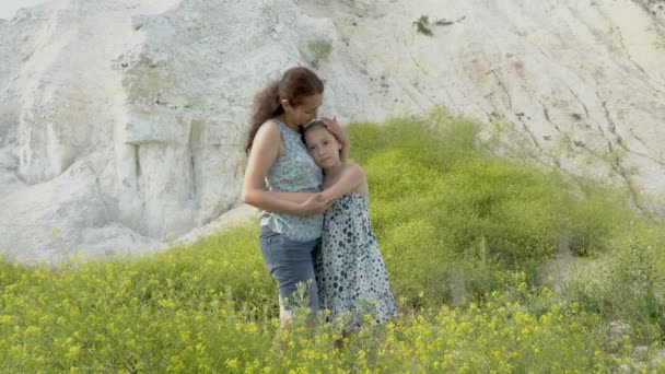 Madre con una hija pequeña en un día soleado contra una montaña de tiza blanca cerca de los arbustos de flores silvestres. Madre gentilmente abraza y besa a su hija. 4K . — Vídeos de Stock