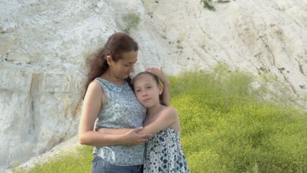 Feliz madre con su pequeña hija en un día soleado contra una montaña de tiza blanca cerca de los arbustos de flores silvestres. Madre gentilmente abraza y besa a su hija. 4K . — Vídeo de stock