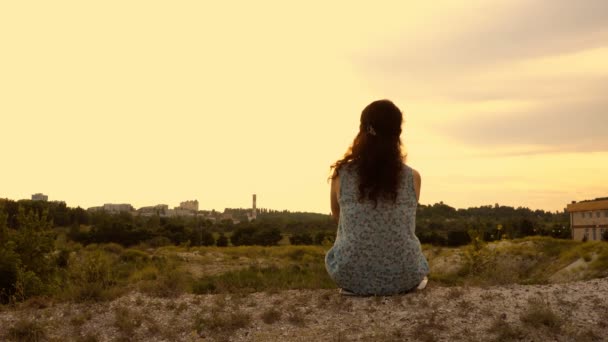 Jovencita al atardecer sentada en la cima de una colina. Ella mira pensativamente a la distancia. Primer plano. 4K — Vídeo de stock