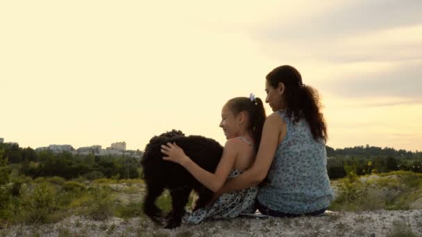 Happy family: mother, daughter and their dog, sitting and playing on top of a hill against a beautiful sunset. They have fun and pet the dog. Close-up. Rear view. The concept of family happiness. 4K — Stock Video