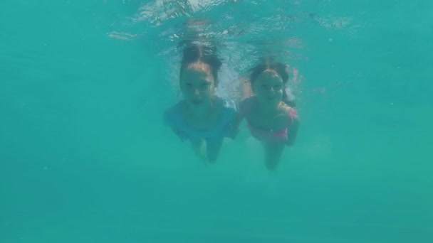 Δύο όμορφα κοριτσάκια κολυμπούν κάτω από το νερό. Κρατούν τα χέρια, χαμογελούν και κοίτα την κάμερα. 4K — Αρχείο Βίντεο