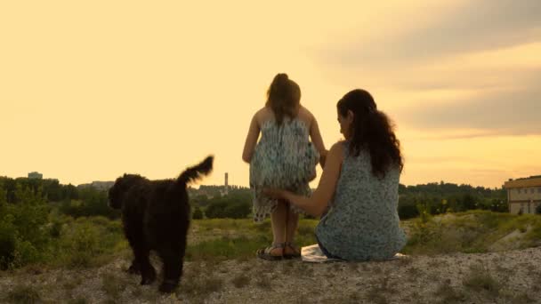 Мать, дочь и их собака сидят на вершине холма против заката и драматического неба. Они гладят собаку и смотрят вдаль. Крупный план. Вид сзади. Понятие семейного счастья. 4K — стоковое видео
