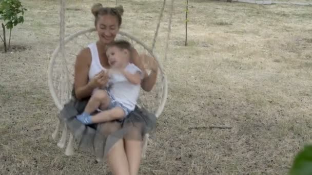 Feliz mamá y el bebé se sientan y columpian en el columpio blanco que cuelgan en el árbol en el parque de la ciudad en un día de verano. Juegan y sonríen. El concepto de felicidad familiar. 4K — Vídeo de stock