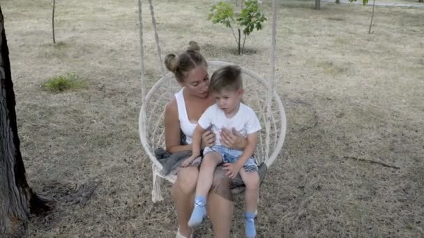 Niño feliz sentado en su regazo de madres en un columpio colgado en un árbol en el parque de la ciudad. Sonríen y ríen. El niño mira a la cámara y agita las manos. 4K — Vídeos de Stock