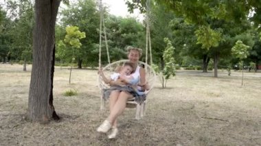 Mutlu anne ve beyaz tişörtlü küçük çocuk şehir Park açık havada bir ağaç asılı bir salıncak üzerinde sallanan vardır. Gülümsüyorlar, oynuyorlar ve eğleniyorlar. 4k