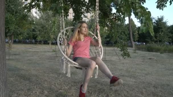 Une jeune fille, blonde tatouée, dans un t-shirt rose, se balançant sur une balançoire suspendue à un arbre dans un parc de la ville à l'extérieur. Elle sourit et se balance sur une chaise en osier blanc. 4K — Video