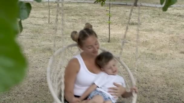 Close-up van moeder en baby die swingend op een witte schommel in de schaduw van een boom in het stads Park op een zomerdag. Het concept van familie geluk. 4k — Stockvideo