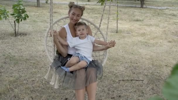 Un petit garçon heureux joue sur les genoux de sa mère, qui est assise sur une balançoire suspendue dans le parc de la ville dans un t-shirt blanc. Ils sourient et rient. Gros plan. 4K . — Video