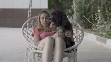 Siyah uzun saçlı köpek oturur ve bir yaz günü bahçesinde beyaz asılı salıncak üzerinde oturan genç bir sarışın kucağında kameraya bakar. Kız sallanan, köpek okşayarak ve gülümseyerek. Close.