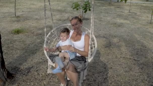 Η μαμά με ένα νεαρό γιο στην αγκαλιά του κάθεται σε μια άσπρη κούνια, η οποία κρέμονται σε ένα δέντρο στο πάρκο το ηλιοβασίλεμα μια καλοκαιρινή μέρα. Χαμογελούν και παίζουν με άσπρα μπλουζάκια. Η θέα από πάνω. 4K. — Αρχείο Βίντεο