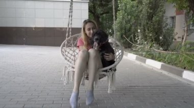 Siyah bir köpek ile birlikte neşeli bir genç sarışın bir yaz günü bahçesinde bir ağaca asılı beyaz bir salıncak üzerinde salıncak. Kız gülümsüyor ve köpeğe sarılıyor. Close. 4k.