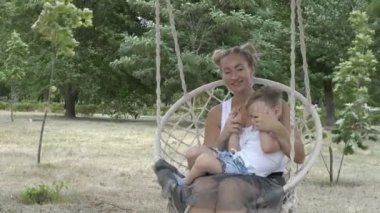 Aile: Anne ve küçük bir çocuk asılı bir salıncak üzerinde Parkta salıncak. Bebek ve anne beyaz t-shirt gülümseme ve yeşil ağaçların arka planda oynamak. Portre. Close. 4k.
