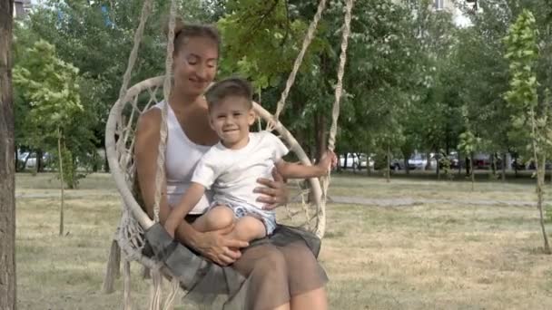 Mãe feliz com o bebê em seu colo sentado e balançando no balanço no parque na árvore. Eles são iluminados pelos raios do sol poente. Bebê e mãe em camisetas brancas sorriem e brincam. Close-up . — Vídeo de Stock