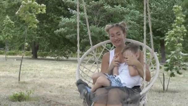Семья: мама с маленьким мальчиком качаются в парке на висячих качелях. Малыш и мама в белых футболках улыбаются и играют на фоне зеленых деревьев. Портрет. Крупный план. 4K . — стоковое видео