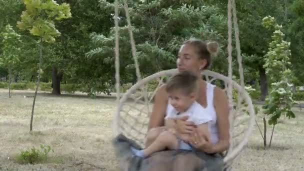 Счастливые мама и ребенок быстро качаются на висячих качелях в парке. Ребенок сидит на коленях у матери, они улыбаются и играют на фоне зеленых деревьев в солнечный день. Крупный план. 4K . — стоковое видео