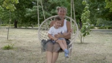 Annesiyle birlikte mutlu bir çocuk, bir yaz gününün gün batımında Park'taki bir ağaca asılan beyaz bir salıncakta sallanıyor. Beyaz tişörtlerle gülümsüyor ve oynuyorlar. Close. 4k.