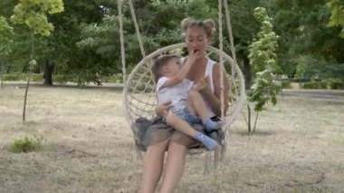 Yakın çekim: mutlu anne ve bebek salıncak ve şehir Park'ta asılı bir salıncak üzerinde oturan oynamak. Çocuk annesinin kucağına oturur, onlar gülümseme ve yeşil bitkilerin arka planda gülmek. Close. 4k.
